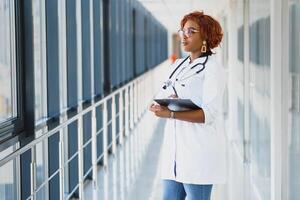 retrato confiante africano americano fêmea médico médico profissional escrevendo paciente notas isolado em hospital clínica corredor janelas fundo. positivo face expressão foto
