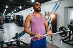 muscular homem fazendo peso elevação exercício às Academia com cópia de espaço. lado Visão do em forma africano americano cara em pé com pesado peso em barra às crossfit clube. maduro homem trabalhando em bíceps às academia. foto