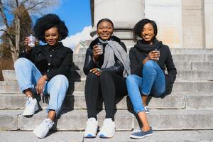 três lindo e à moda pele escura meninas com grandes cabelo em pé dentro uma cidade e bebendo uma café e usar a telefones foto