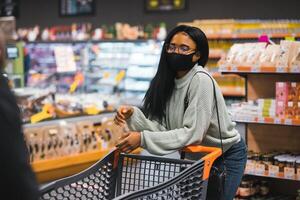 africano mulher vestindo descartável médico mascarar compras dentro supermercado durante coronavírus pandemia surto. epidemia Tempo foto