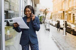 franzindo a testa preocupado Gerente Falando em celular fora. jovem africano americano o negócio mulher em pé perto ao ar livre vidro muro. Móvel telefone conversa conceito foto