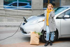 tendo uma conversa usando o telefone. mulher na estação de carga de carros elétricos durante o dia. veículo novo foto