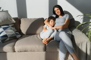 alegre mãe sentado em sofá dentro acolhedor luz vivo quarto detém em colo cócegas do pequeno filho apreciar engraçado Atividades em final de semana às lar, ter Diversão junto, impagável Tempo com pré escola criança conceito foto