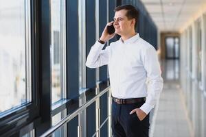 Visão dentro perfil do uma jovem homem de negocios, vestido dentro uma branco camisa em pé perto a janela em uma azul escuro paredes e olhando Fora a janela. foto