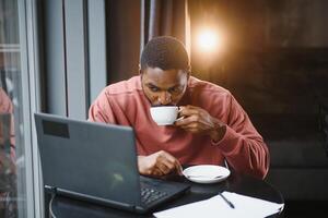 retrato do feliz africano homem de negocios usando telefone enquanto trabalhando em computador portátil dentro restaurante. foto