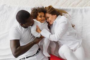 retrato do feliz multirracial jovem família deitado em acolhedor branco cama às lar, sorridente internacional mãe e Papai relaxante com pequeno birracial menina criança posando para cenário dentro quarto foto