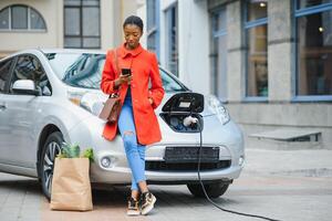 usando Smartphone enquanto esperando. africano americano menina em a elétrico carros carregar estação às dia. marca Novo veículo foto