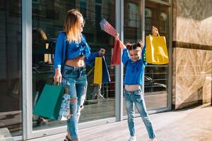 lindo mãe e dela fofa pequeno filha estão segurando compras bolsas, olhando às Câmera e sorridente enquanto em pé ao ar livre. compras conceito foto