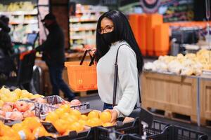 africano mulher vestindo descartável médico mascarar. compras dentro supermercado durante coronavírus pandemia surto. epidemia Tempo foto