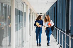 dois jovem fêmea alunos em pé com livros e bolsas dentro a corredor universidade Falando cada outro. foto