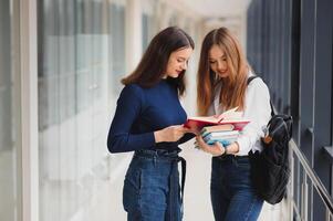 dois jovem fêmea alunos em pé com livros e bolsas dentro a corredor universidade Falando cada outro. foto