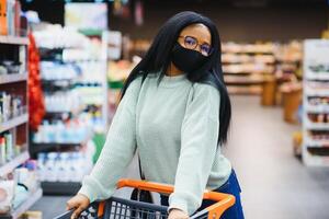 africano mulher vestindo descartável médico mascarar. compras dentro supermercado durante coronavírus pandemia surto. epidemia Tempo foto