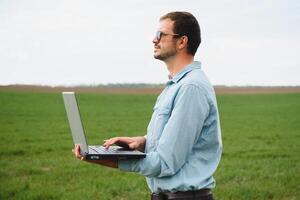 homem agricultor trabalhando em uma computador portátil dentro a campo. agrônomo examina a verde brotar inverno trigo. foto