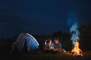 jovem casal tendo uma descansar às fogueira ao lado acampamento e azul turista barraca, bebendo chá, desfrutando noite céu. a conceito do ativo lazer e viagem com uma barraca foto
