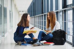 dois bonita fêmea alunos com livros sentado em a chão dentro a universidade corredor foto