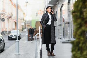 atraente jovem mulher dentro a cidade. o negócio senhora é sorridente enquanto falando em uma inteligente telefone dentro frente do moderno o negócio Centro foto