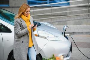 usando smartphone enquanto espera. mulher na estação de carga de carros elétricos durante o dia. veículo novo foto