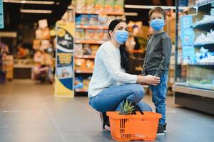 mãe e dela filho vestindo protetora face mascarar fazer compras às uma supermercado durante a coronavírus epidemia. foto