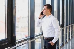 retrato do uma sério confiante homem patrão tendo Móvel telefone conversação enquanto em repouso depois de encontro com dele parceiros, homem de negocios falando em célula Telefone enquanto em pé dentro moderno espaço dentro de casa foto