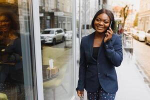 franzindo a testa preocupado Gerente Falando em celular fora. jovem africano americano o negócio mulher em pé perto ao ar livre vidro muro. Móvel telefone conversa conceito foto