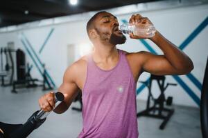 jovem africano americano desportista bebendo água e olhando longe dentro academia. foto