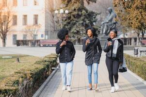 três lindo e à moda pele escura meninas com grandes cabelo em pé dentro uma cidade e bebendo uma café e usar a telefones foto