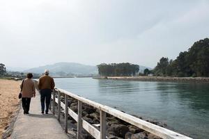 casal de 70-80 anos andando em rodiles, visto de costas. caminho de madeira próximo ao rio com puntal ao fundo foto