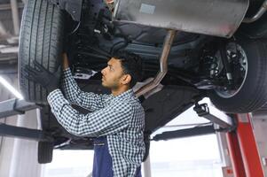 indiano carro mecânico em pé e trabalhando dentro serviço estação. carro especialistas examinando a levantado carro. profissional reparadores vestindo mecânico uniforme dentro azul cor. foto