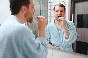 escovar dentes dentro a manhã. traseiro Visão do bonito jovem barba homem escovar dele dentes e sorridente enquanto em pé contra uma espelho. foto