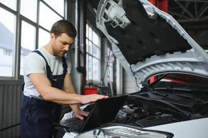 mecânico homem mecânico Gerente trabalhador usando uma computador portátil computador verificação carro dentro oficina às auto carro reparar serviço Centro. engenheiro jovem homem olhando às inspeção veículo detalhes debaixo carro de capuz foto