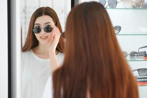 retrato do uma jovem mulher compras, em pé dentro loja e tentando oculos de sol perto uma espelho. foto