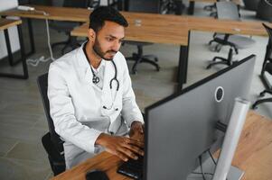 pessoas, ocupação e remédio conceito. sorridente masculino indiano árabe médico dentro branco casaco, sentado dentro médico escritório às escrivaninha com computador portátil foto