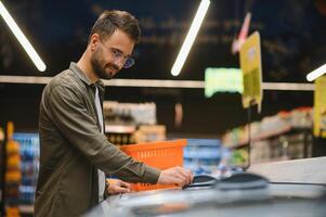 jovem homem comprando mercearias às a supermercado. de outros clientes dentro fundo. consumismo conceito. foto