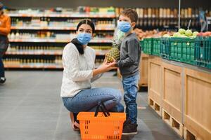 jovem mulher e dela criança vestindo protetora face máscaras fazer compras uma Comida às uma supermercado durante a coronavírus epidemia ou gripe surto. foto
