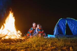 jovem casal tendo uma descansar às fogueira ao lado acampamento e azul turista barraca, bebendo chá, desfrutando noite céu. a conceito do ativo lazer e viagem com uma barraca foto