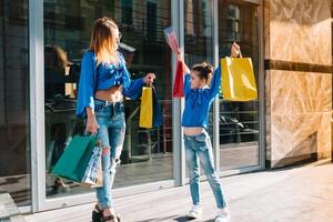 lindo mãe e dela fofa pequeno filha estão segurando compras bolsas, olhando às Câmera e sorridente enquanto em pé ao ar livre. compras conceito foto