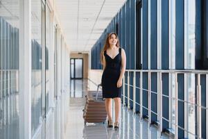 à moda mulher dentro uma Preto vestir com uma mala de viagem dentro a aeroporto terminal esperando para uma avião. foto