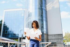 retrato do jovem fêmea africano americano trabalho buscador guardando uma pasta com cv dentro dela mãos em pé contra escritório prédio. borrado fundo com cópia de espaço foto