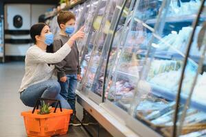 compras com criança durante vírus surto. mãe e criança vestindo cirúrgico face mascarar comprando fruta dentro supermercado. mãe e pequeno Garoto Comprar fresco vegetal dentro mercearia loja. família dentro fazer compras foto