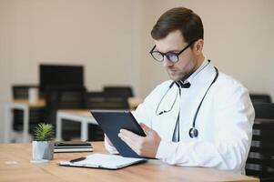 a moderno doutor. cortada tiro do uma bonito masculino médico trabalhando em dele tábua enquanto sentado dentro dele escritório. foto