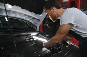 bonito jovem masculino auto mecânico dentro especial uniforme roupas segurando uma lanterna, olhando para demolir e reparação debaixo a de capuz dentro a carro motor dentro uma carro oficina foto