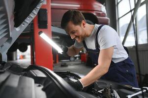 bonito jovem masculino auto mecânico dentro especial uniforme roupas segurando uma lanterna, olhando para demolir e reparação debaixo a de capuz dentro a carro motor dentro uma carro oficina foto