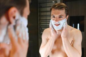 jovem sorridente caucasiano sem camisa homem aplicando barbear espuma em face dentro frente do espelho, preparando para cabelo remoção manhã rotina. conceito do pessoal higiene, cuidados com a pele, masculino facial beleza produtos. foto