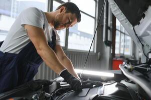 bonito jovem masculino auto mecânico dentro especial uniforme roupas segurando uma lanterna, olhando para demolir e reparação debaixo a de capuz dentro a carro motor dentro uma carro oficina foto