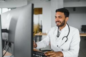 pessoas, ocupação e remédio conceito. sorridente masculino indiano árabe médico dentro branco casaco, sentado dentro médico escritório às escrivaninha com computador portátil foto
