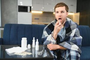 rinite, remédio e cuidados de saúde conceito - doente homem dentro cobertor usando oral spray às lar. foto