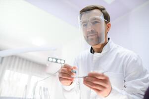 inferior Visão jovem confiante feliz profissional estomatologista europeu médico homem 20s dentro branco vestido aguarde mostrando dente perfuração máquina às dentista escritório interior gabinete. cuidados de saúde estético tratamento. foto