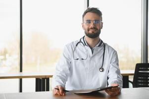 médico homem sentado às a escrivaninha às dele trabalhando lugar. perfeito médico serviço dentro clínica. retrato do uma bonito jovem doutor. foto