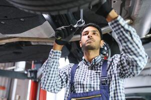 indiano carro mecânico em pé e trabalhando dentro serviço estação. carro especialistas examinando a levantado carro. profissional reparadores vestindo mecânico uniforme dentro azul cor. foto