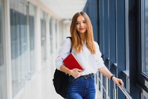 alegre morena aluna menina com Preto mochila detém livros dentro moderno prédio. fêmea aluna em pé com livros dentro Faculdade corredor foto